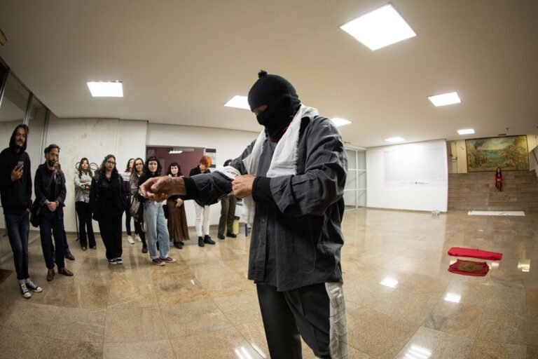 O performer encapuzado amarra uma tira de pano no saguão, com o público de pé em roda e panos vermelhos com terra no chão