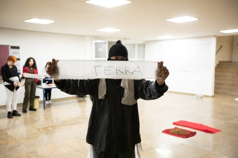 O performer encapuzado ergue uma tira de pano com a palavra "terra", com panos vermelhos com terra no chão