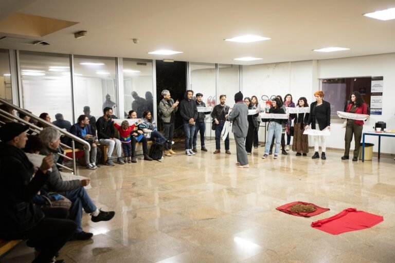 O público forma uma roda no saguão da Pinacoteca segurando as tiras de pano, com os panos vermelhos no centro
