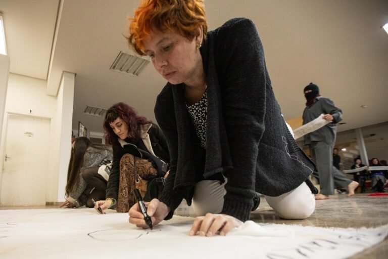 A mulher ruiva escreve no pano branco enquanto o performer encapuzado passa ao fundo com uma tira de pano
