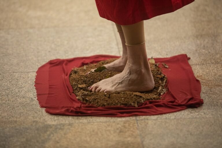 Detalhe dos pés descalços da performer, de pé sobre terra num pano vermelho