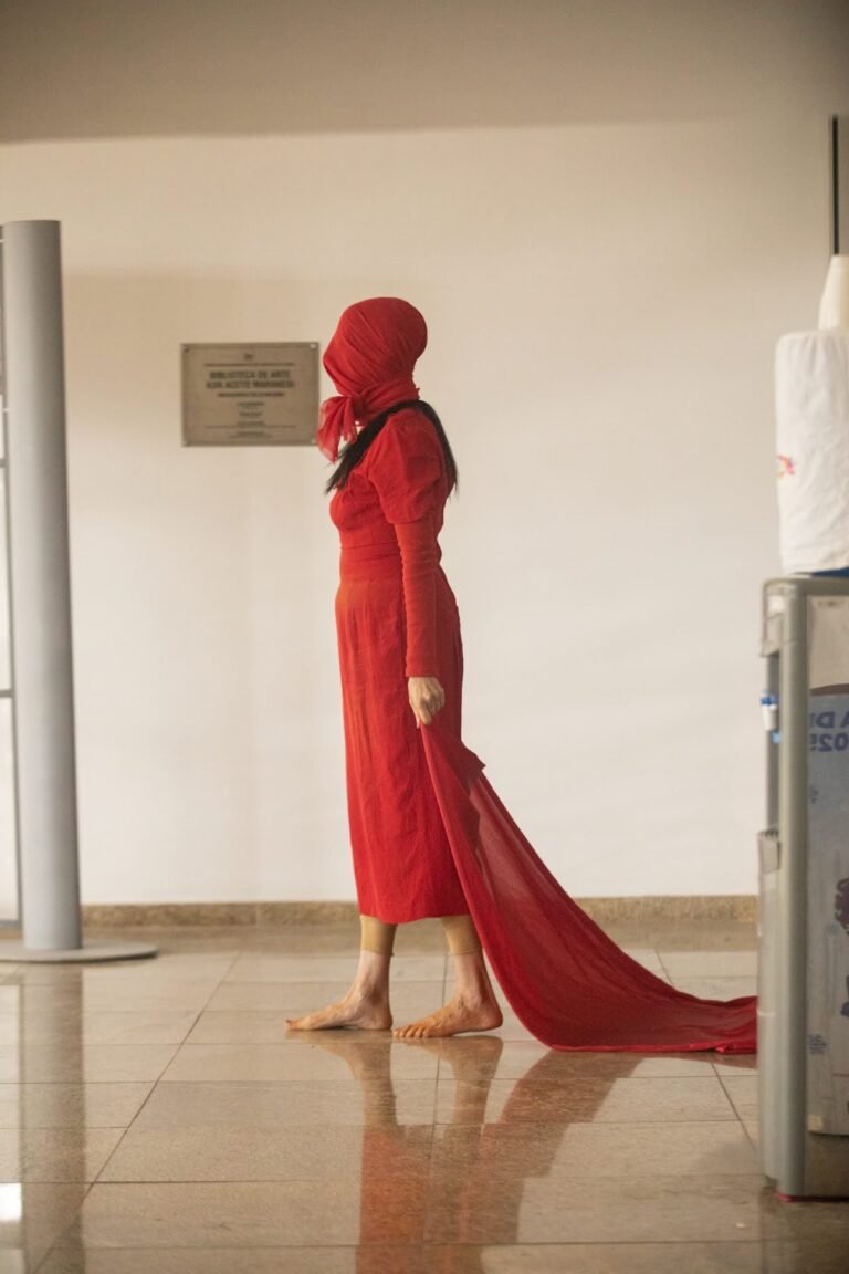 A performer, coberta de vermelho da cabeça aos pés, arrasta uma longa cauda de tecido vermelho pelo saguão