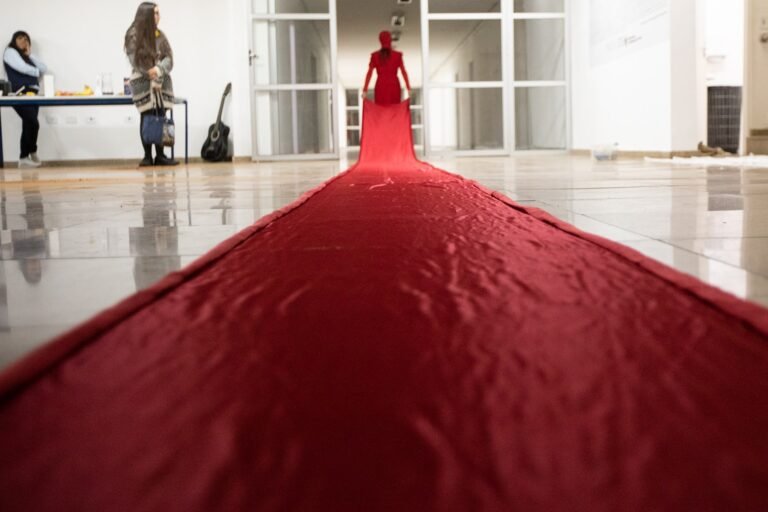Longa cauda de tecido vermelho estendida no chão do saguão, com a performer ao fundo
