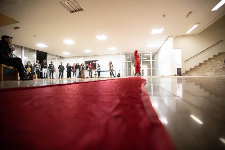 A cauda vermelha atravessa o saguão até a performer, com o público de pé ao longo das paredes
