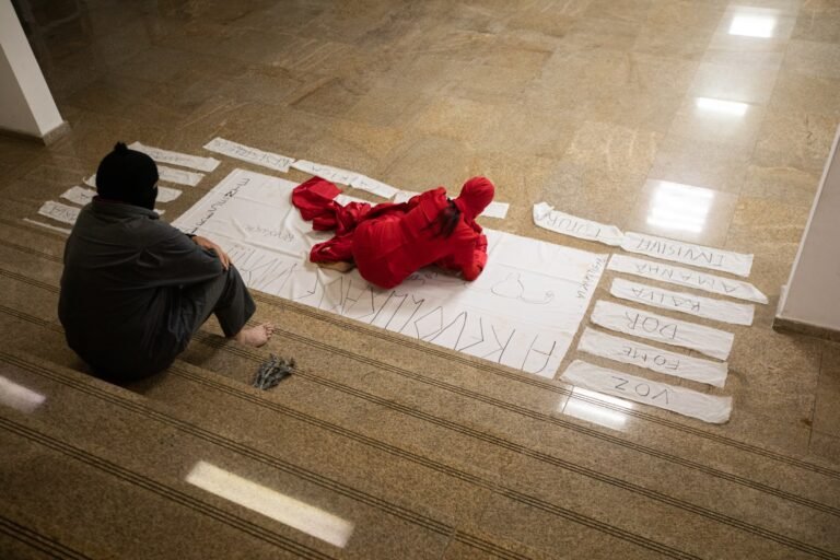 A performer deitada sobre o pano branco cercado pelas tiras de palavras, observada pelo performer encapuzado sentado na escada