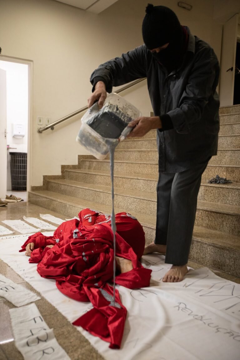 O performer encapuzado despeja o líquido cinza do balde sobre o corpo envolto em vermelho