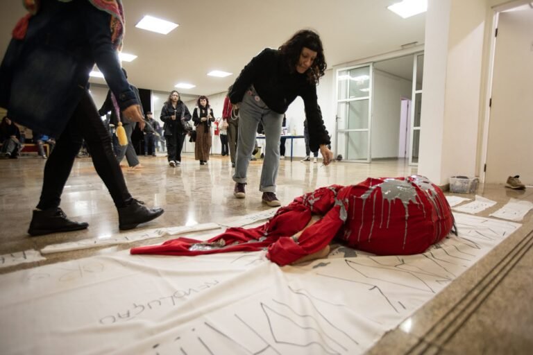 Pessoas do público caminham ao redor do corpo coberto de tecido vermelho e tinta cinza sobre o pano branco