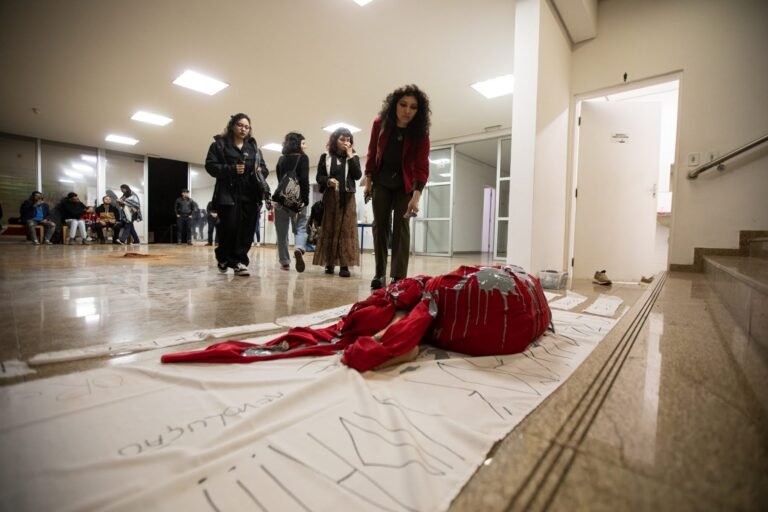 Pessoas do público se aproximam do corpo envolto em vermelho e cinza, estendido sobre o pano branco no saguão