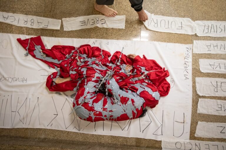Vista de cima do corpo envolto em tecido vermelho, coberto de tinta cinza e flores secas, sobre o pano branco escrito e cercado por tiras com palavras como "fábrica", "greve" e "futuro"