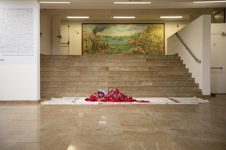 O corpo envolto em vermelho, sobre o pano branco, ao pé da escadaria vazia do saguão da Pinacoteca, sob o grande painel pintado