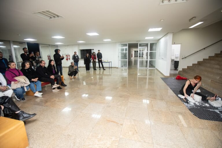 Kareen Labbé Weber ajoelhada sobre uma tela no canto do saguão da Pinacoteca, com o público sentado assistindo