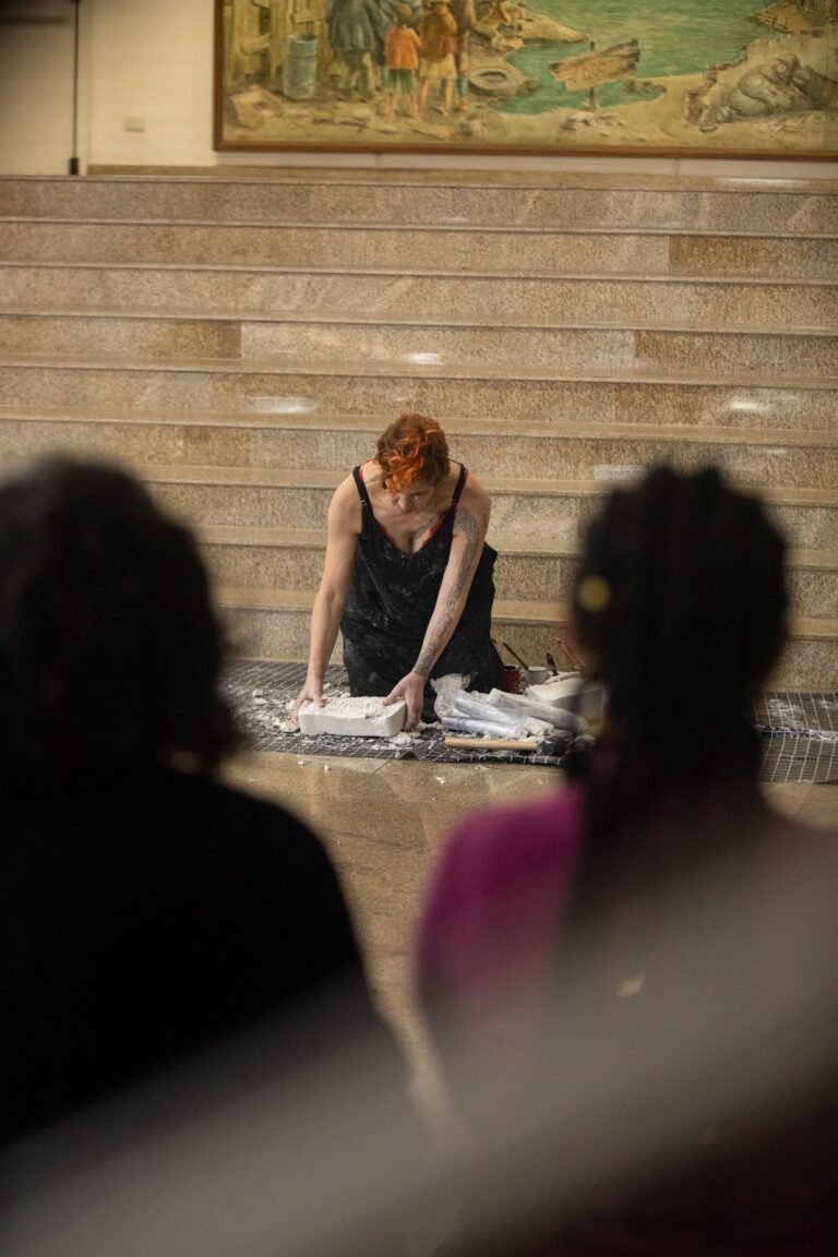 Kareen Labbé Weber, de vestido preto, curvada sobre os blocos de gesso ao pé da escadaria, vista entre duas pessoas do público