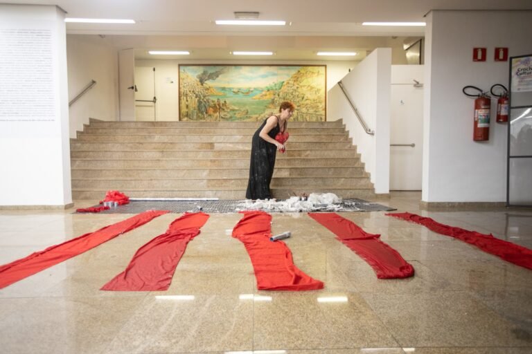 Faixas de tecido vermelho estendidas no chão do saguão, com Kareen Labbé Weber ao pé da escadaria, sob o grande painel pintado