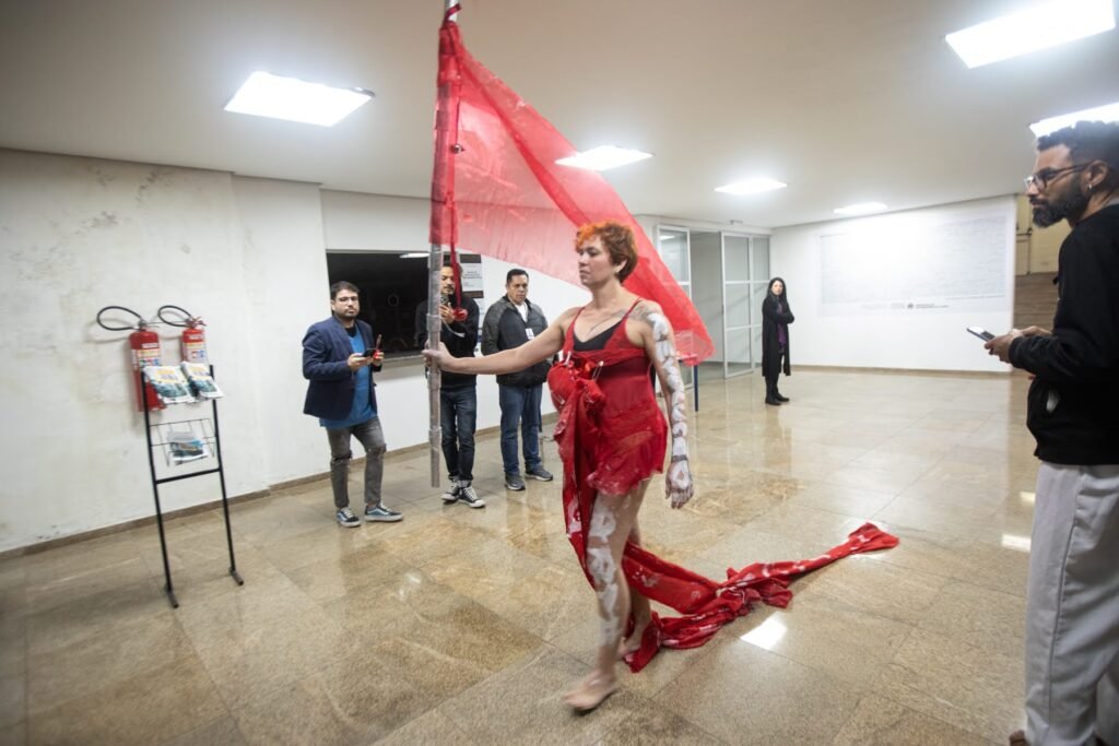 Kareen Labbé Weber caminha pelo saguão com uma bandeira vermelha e a cauda do vestido arrastando no chão