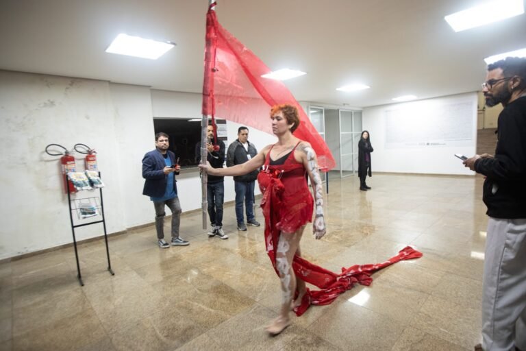 Kareen Labbé Weber caminha pelo saguão com uma bandeira vermelha e a cauda do vestido arrastando no chão