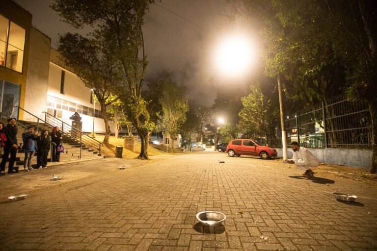 PC agachado no meio de bacias de alumínio espalhadas pela praça, com o público junto ao prédio