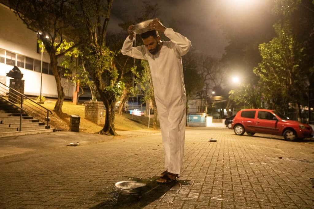 PC ergue uma bacia sobre a cabeça na praça iluminada por um poste, com um carro vermelho ao fundo