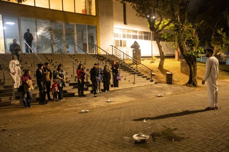 PC de pé na praça, diante do público reunido na escadaria da Pinacoteca, com bacias espalhadas pelo chão