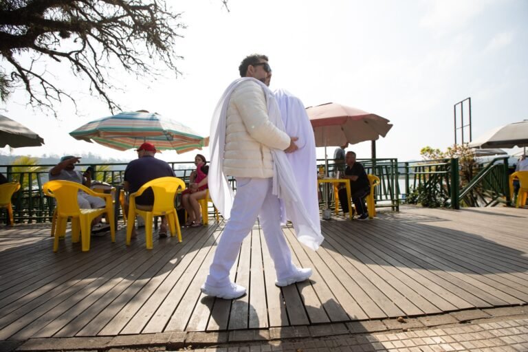 Pio Santana, todo de branco, caminha no deck de um restaurante à beira da Represa Billings carregando tecidos brancos