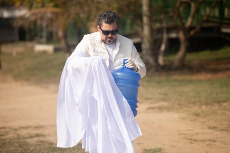 Pio Santana segura um galão de água azul junto ao corpo, entre tecidos brancos