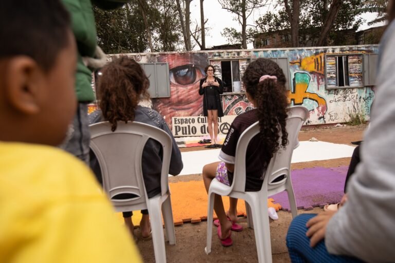 Reya Perovan fala ao público sentado em cadeiras de plástico no pátio do Espaço Cultural Pulo do Sapo
