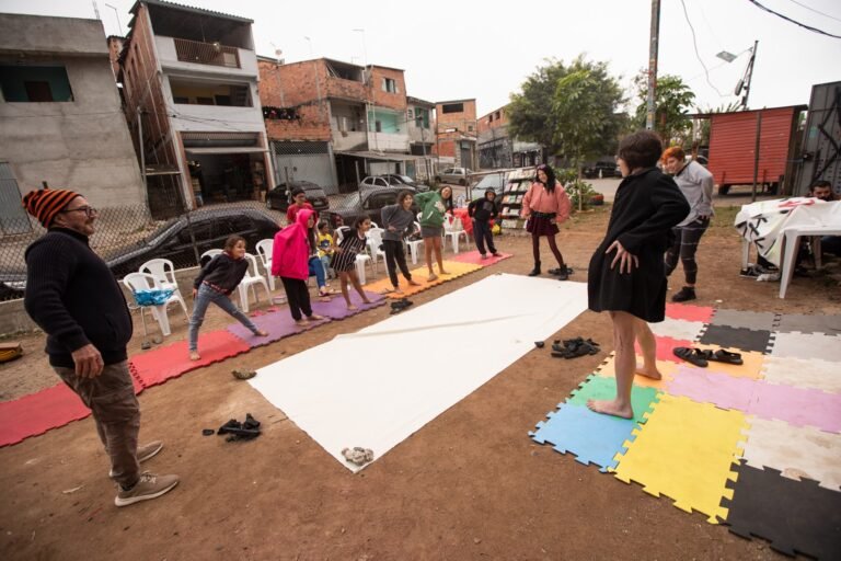 Participantes ao redor do longo tecido cru estendido sobre tapetes coloridos no pátio, com as casas do bairro ao fundo