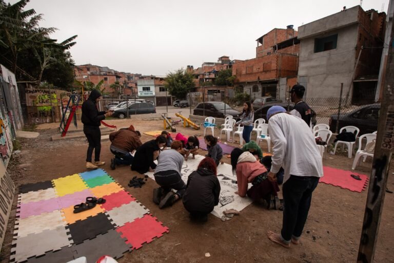 Participantes ajoelhados desenham juntos no tecido cru, no pátio do Pulo do Sapo, com as casas do bairro ao fundo