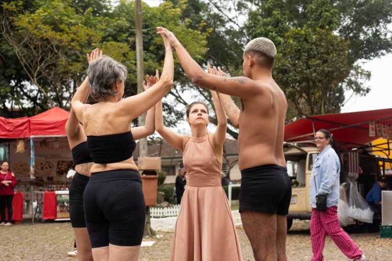 Performers em roda, com as mãos unidas no alto, entre as barracas da feira