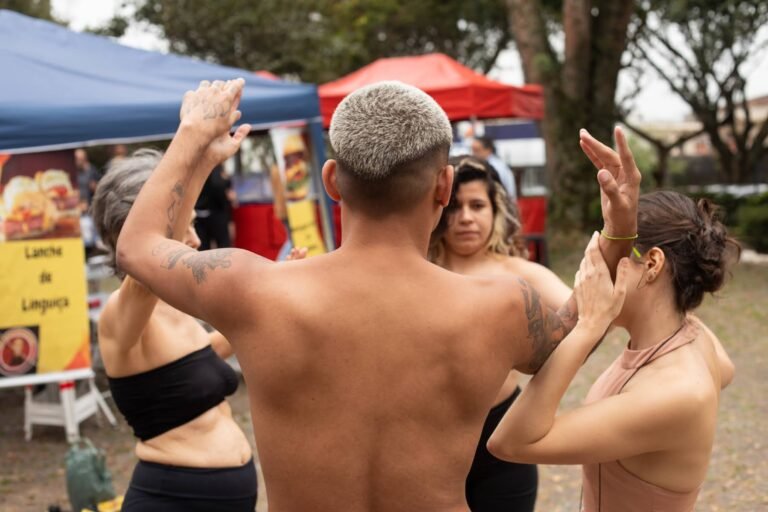 Um performer de cabelo platinado, de costas, ergue os braços entre as performers