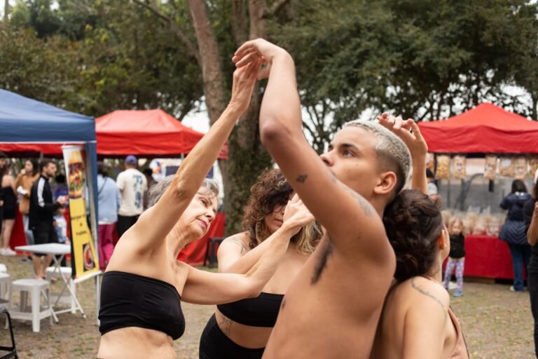 As performers se entrelaçam de braços erguidos, com barracas vermelhas ao fundo