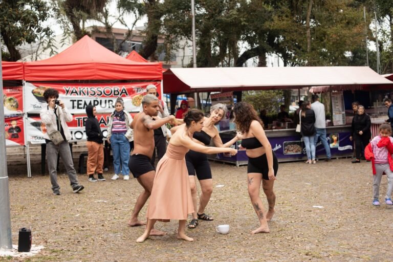 As quatro performers dançam na praça de terra, observadas pelo público da feira