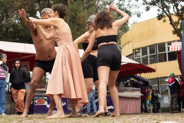 As performers se movem juntas, uma delas de vestido rosado, diante das barracas e de um prédio amarelo