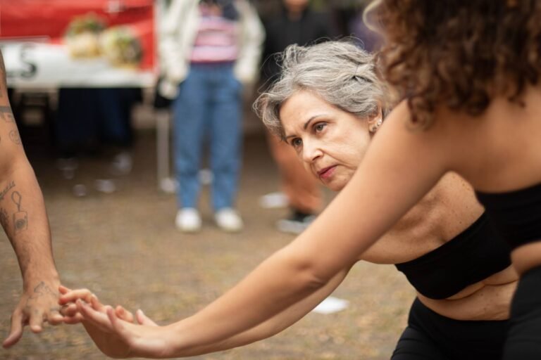 Uma performer de cabelos grisalhos estende o braço e toca a mão de outra performer
