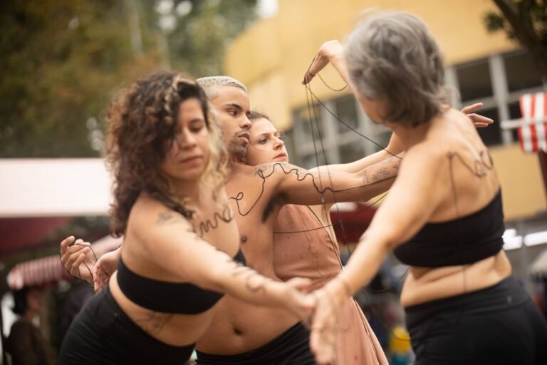 As performers dançam entrelaçadas, a linha preta escrevendo palavras sobre os corpos