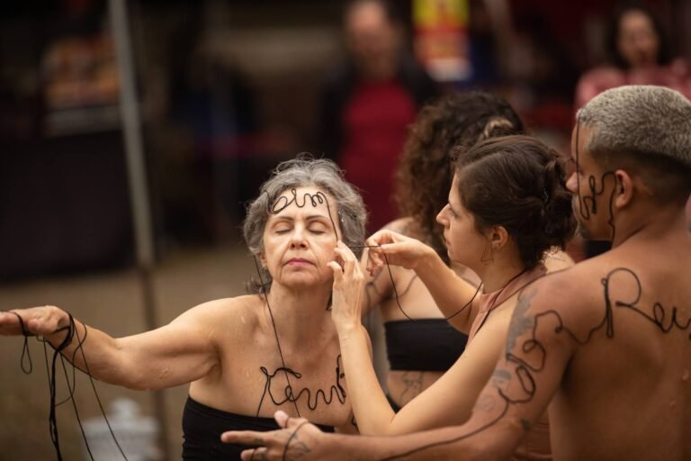A performer de cabelos grisalhos, de olhos fechados, com a linha preta sobre a testa, cercada pelas outras