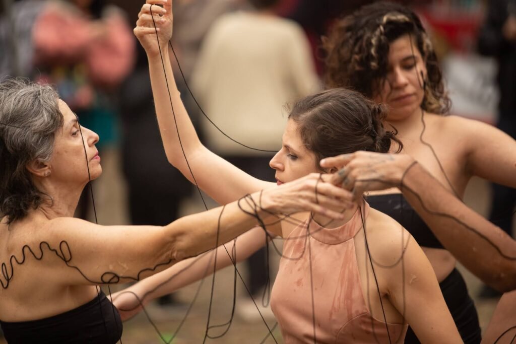 As performers enrolam a linha preta no pescoço e nos braços de uma delas