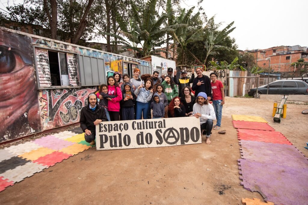Um grupo de pessoas posa sorridente para a foto atrás de uma placa retangular grande com o texto Espaço Cultural Pulo do Sapo. A letra A de sapo é envolta em um círculo remetendo ao símbolo para anarquia. O grupo é formado por diversas crianças e adultos. Há um conteiner com janela, o chão é de terra com tapetes de E.V.A.. Ao fundo, casas com tijolo aparente vermelho e algumas árvores.