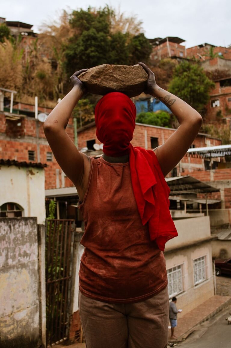 Dani Mara, de costas e com capuz vermelho, equilibra uma pedra sobre a cabeça diante de casas de tijolo