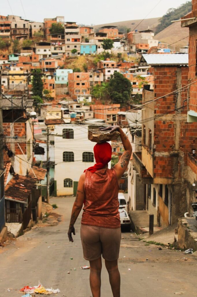 Dani Mara, de costas, sobe uma rua de bairro carregando uma pedra sobre a cabeça