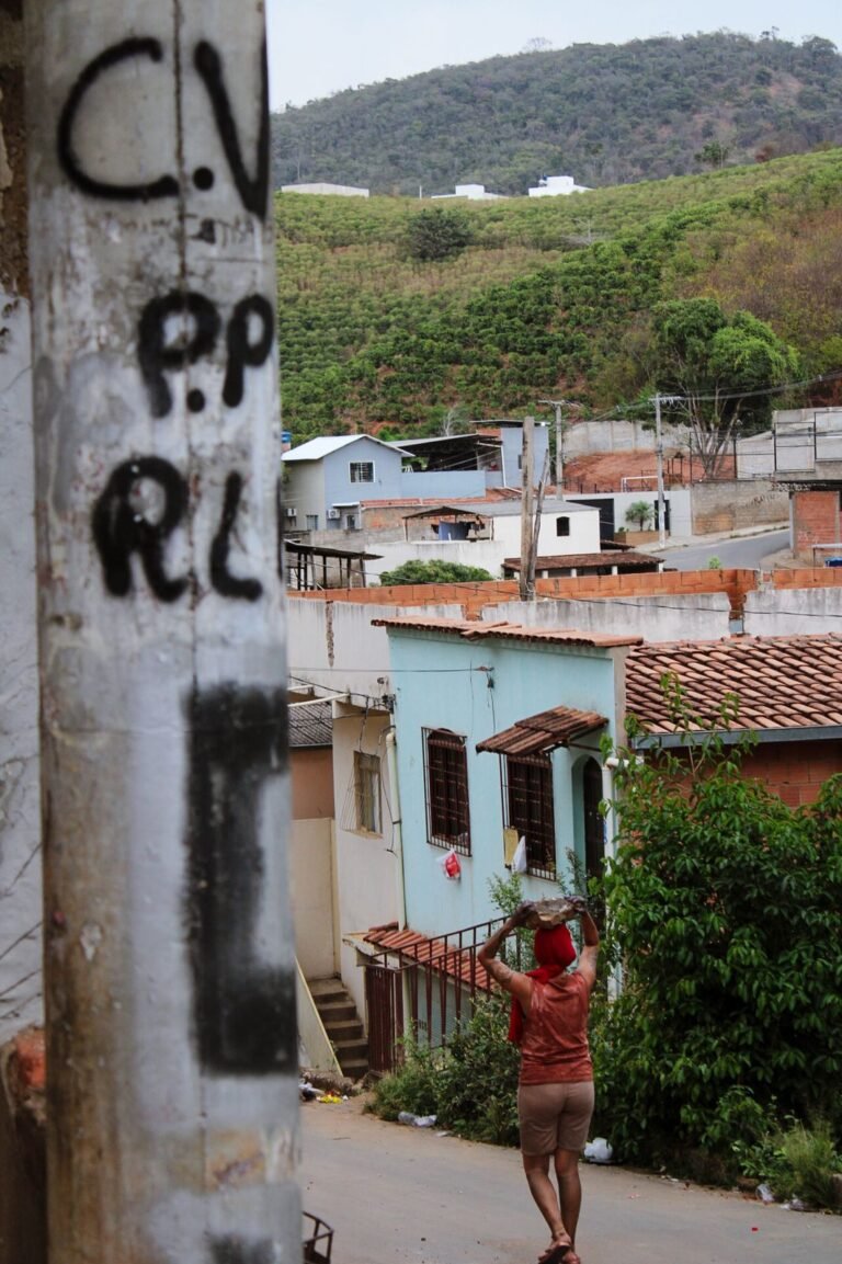 Rua de bairro vista ao lado de um poste pichado; ao fundo, Dani Mara caminha com uma pedra na cabeça