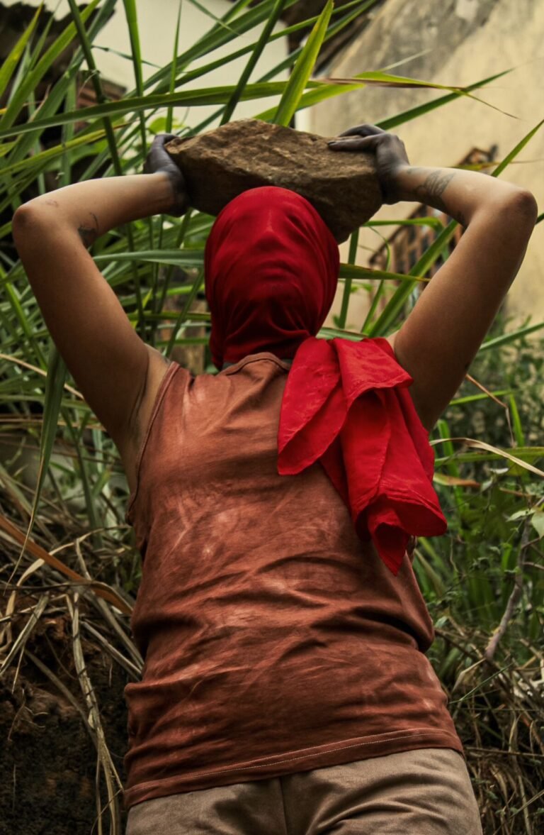 Close de Dani Mara, de costas, segurando com as duas mãos uma pedra sobre a cabeça coberta por um tecido vermelho