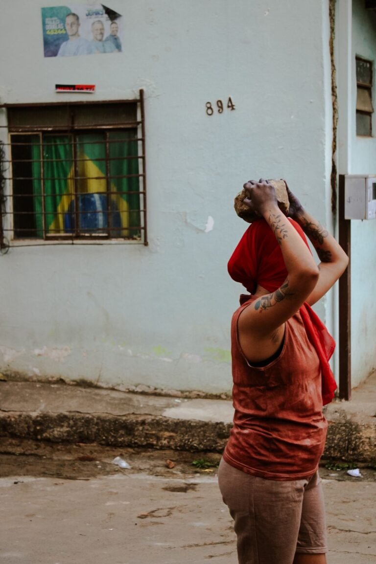 Dani Mara, de perfil, segura uma pedra sobre a cabeça em frente a uma casa azul